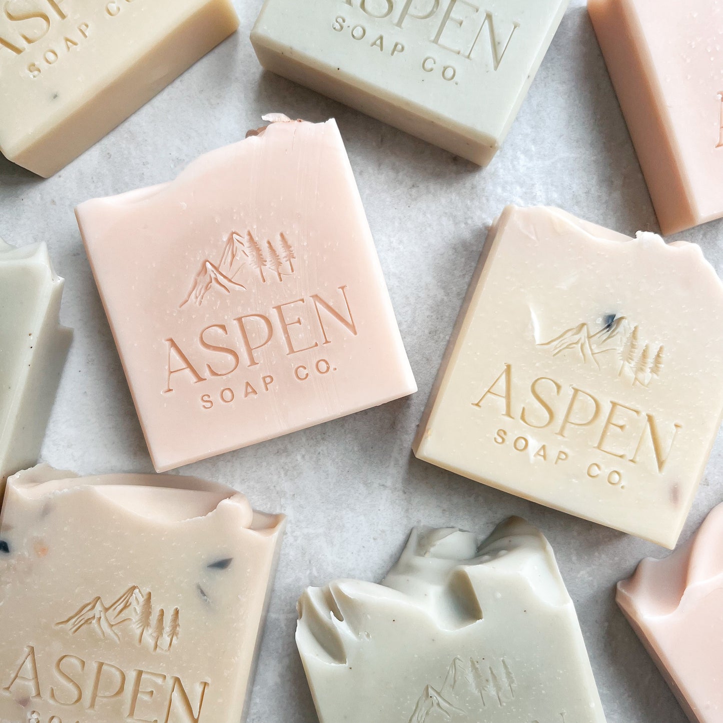 A flat lay of soaps with no label. Each soap is stamped with the Aspen Soap Company logo. Soaps are light pink, light green, and ivory with speckles.
